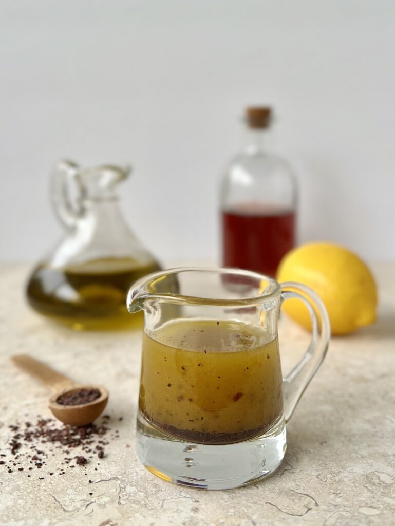 The dressing is pictured in a glass cup with a handle and a lip to pour. In the background are a container of olive oil, vinegar and a lemon. To the side is a wooden teaspoon of sumac.