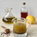 The dressing is pictured in a glass cup with a handle and a lip to pour. In the background are a container of olive oil, vinegar and a lemon. To the side is a wooden teaspoon of sumac.