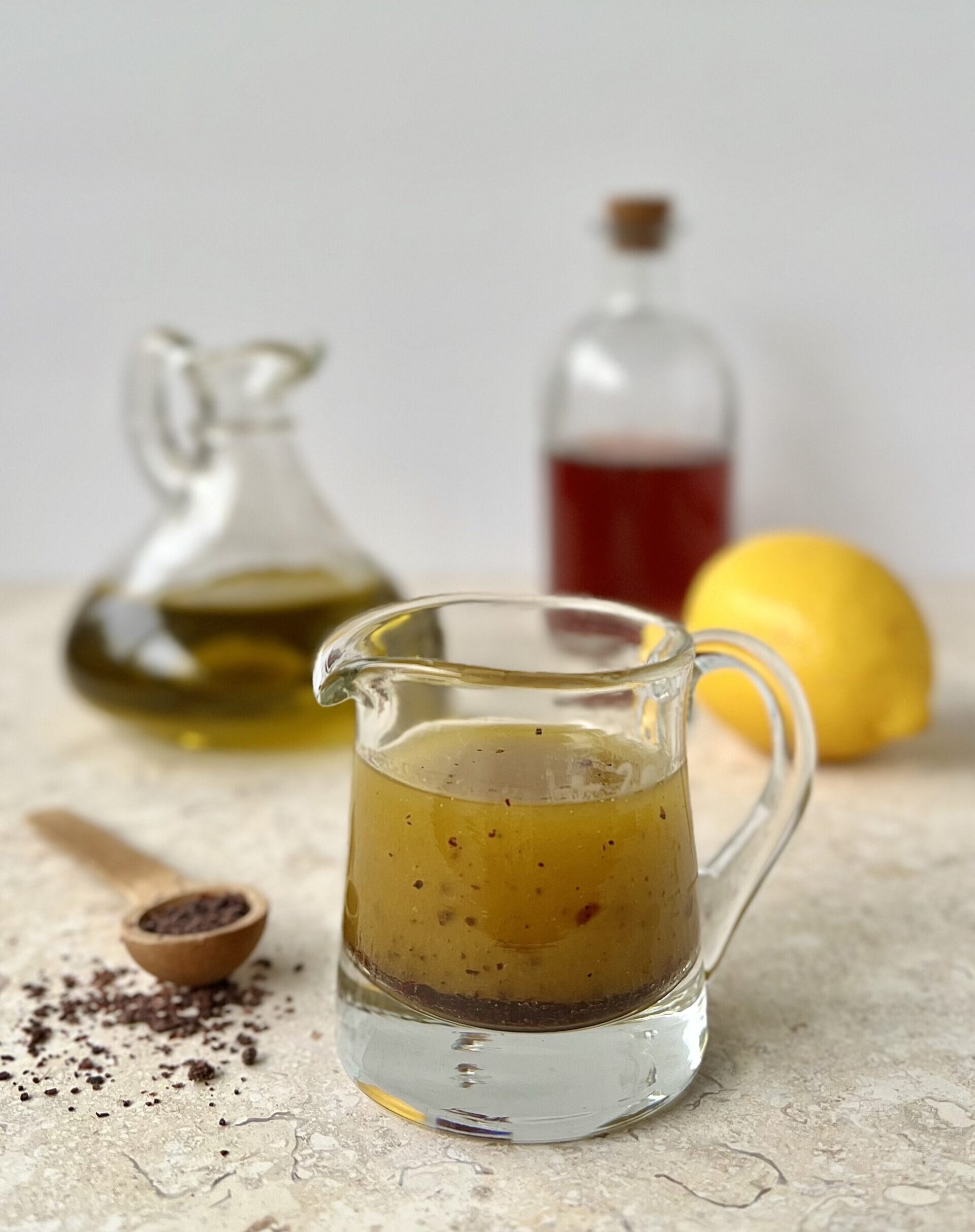 The dressing is pictured in a glass cup with a handle and a lip to pour. In the background are a container of olive oil, vinegar and a lemon. To the side is a wooden teaspoon of sumac.