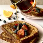 2 pieces of french toast with syrup being actively poured over top of them. There are berries on the french toast.