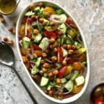 Overhead view of a chopped salad in a white serving dish. Colors of tomatoes, cucumber and chickpeas. There is a spoon on the left side for added effect.