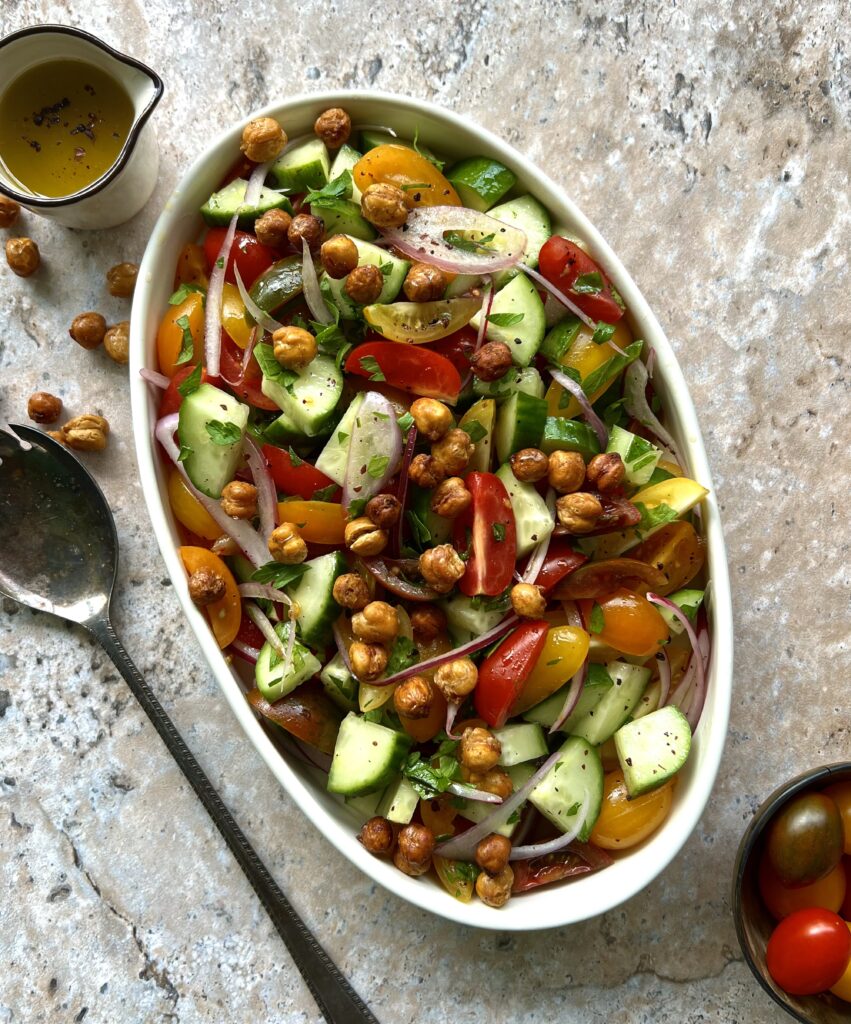 Overhead view of a chopped salad in a white serving dish. Colors of tomatoes, cucumber and chickpeas. There is a spoon on the left side for added effect.