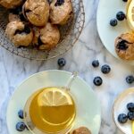 Overhead view of a basket of blueberry muffins, a cup of eat with lemon and a white marble background.