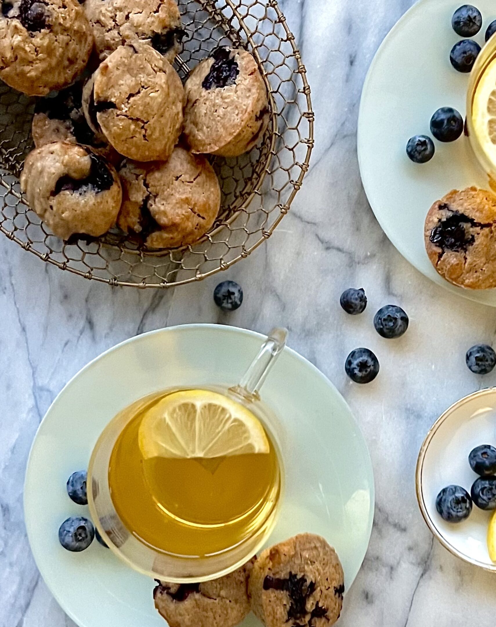 Overhead view of a basket of blueberry muffins, a cup of eat with lemon and a white marble background.