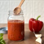 Side view of a glass jar with red pepper sauce inside of it. There is a wooden spoon reaching into the jar. A red pepper and garlic clove sit to the side