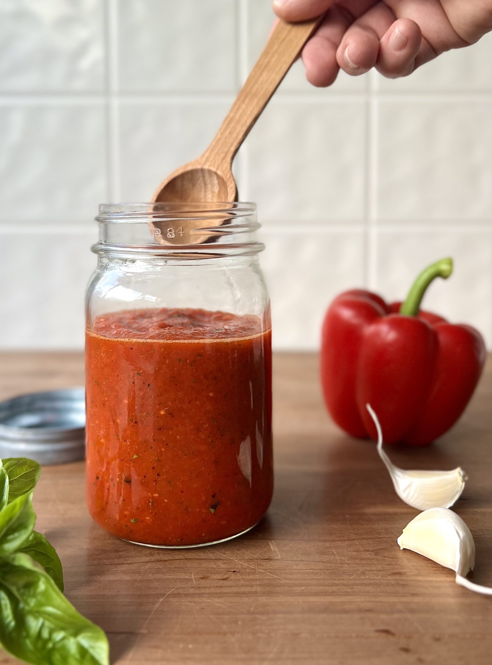 Side view of a glass jar with red pepper sauce inside of it. There is a wooden spoon reaching into the jar. A red pepper and garlic clove sit to the side