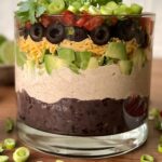 Sideways view of the bean dip in a glass jar. You can see each layer clearly. It is on a wooden cutting board.