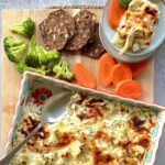 Overhead view of artichoke dip in a colorful baking dish. It is sitting on a brown cutting board that has carrots, bread clices and broccoli on it.