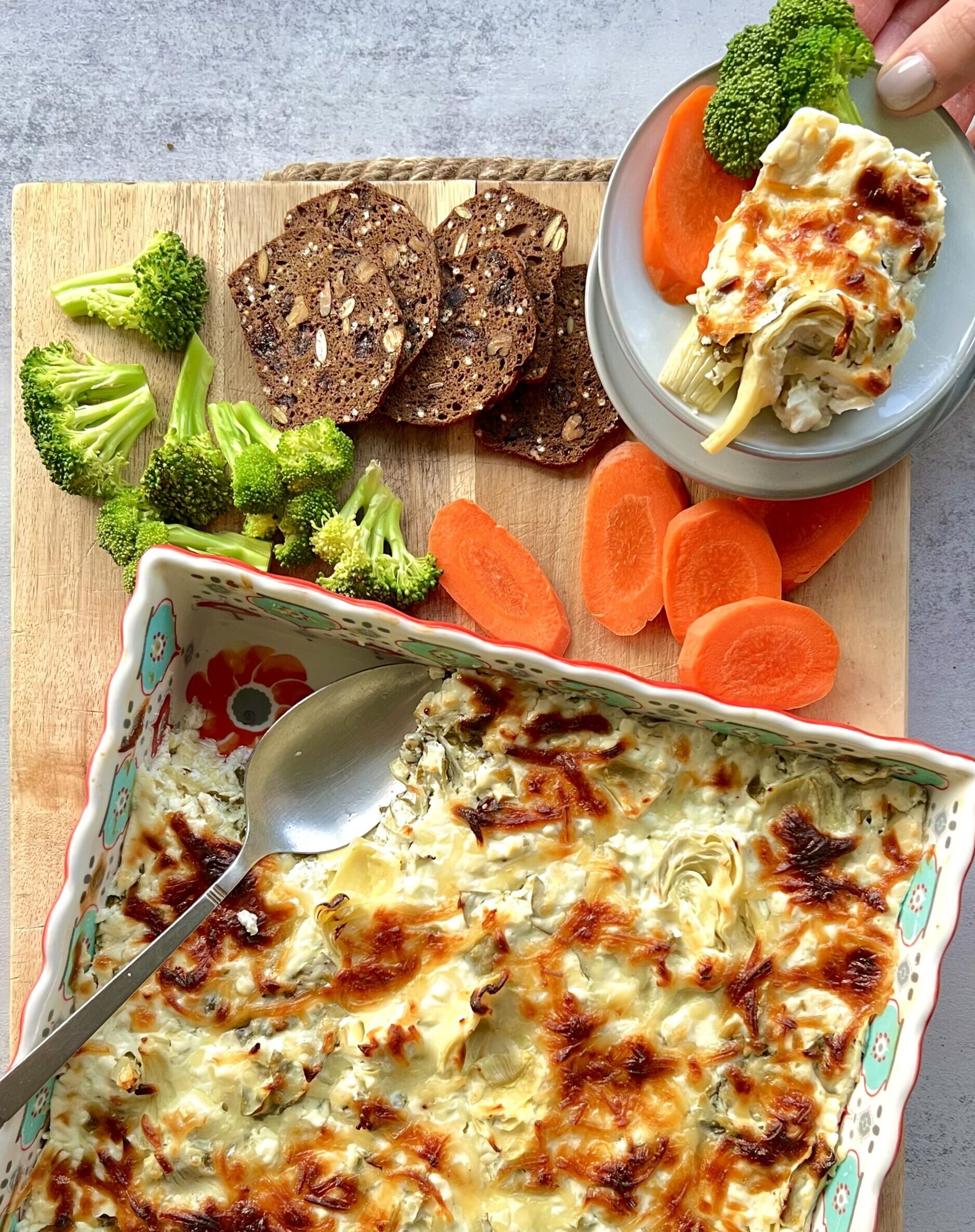 Overhead view of artichoke dip in a colorful baking dish. It is sitting on a brown cutting board that has carrots, bread clices and broccoli on it.