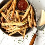 Overhead view of a bowl of air fryer french fries with parmesean cheese and herbs sprinkled on top. At the top, there is a side of ketchup.