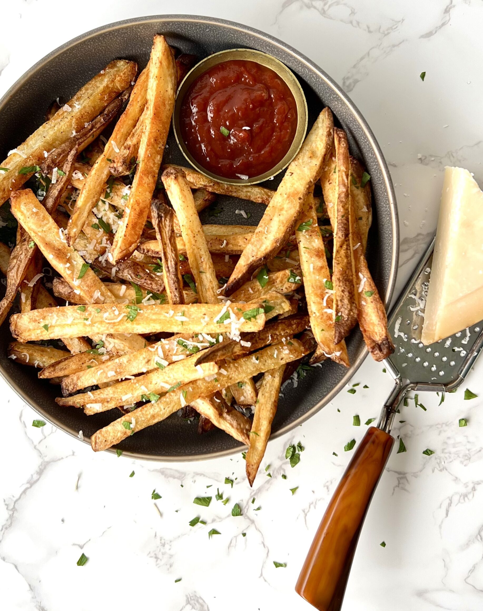 Overhead view of a bowl of air fryer french fries with parmesean cheese and herbs sprinkled on top. At the top, there is a side of ketchup.