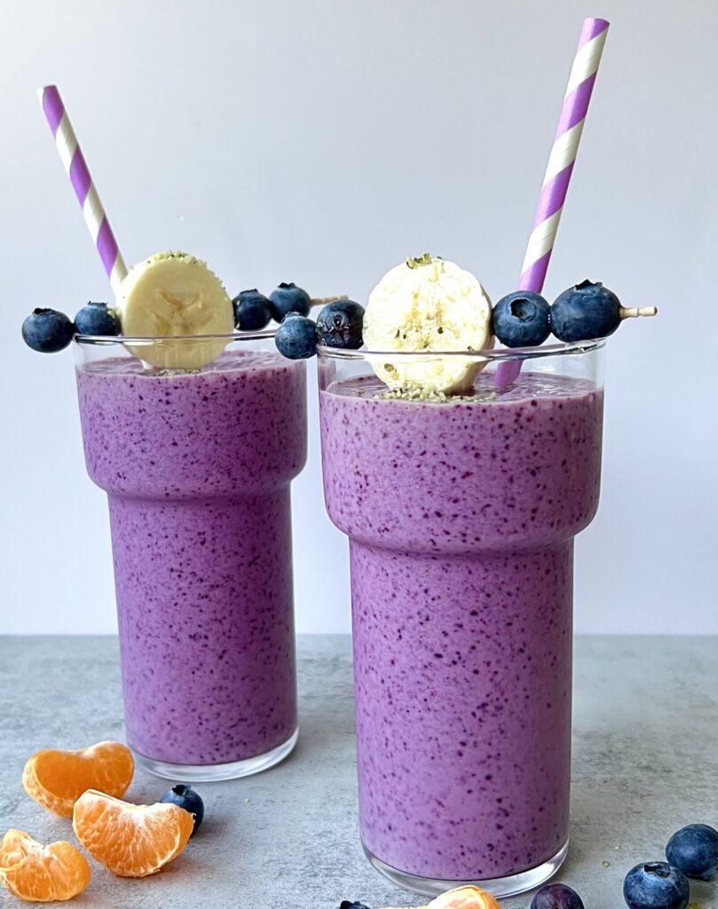 Side view of two beautiful purple smoothies in glasses with slices of banana and blueberries on top. There is a Purple and white straw in each of them.