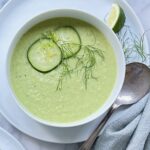 Overhead view of a green gazpacho in a white bowl, garnished by fennel fronds and cucumber slices.