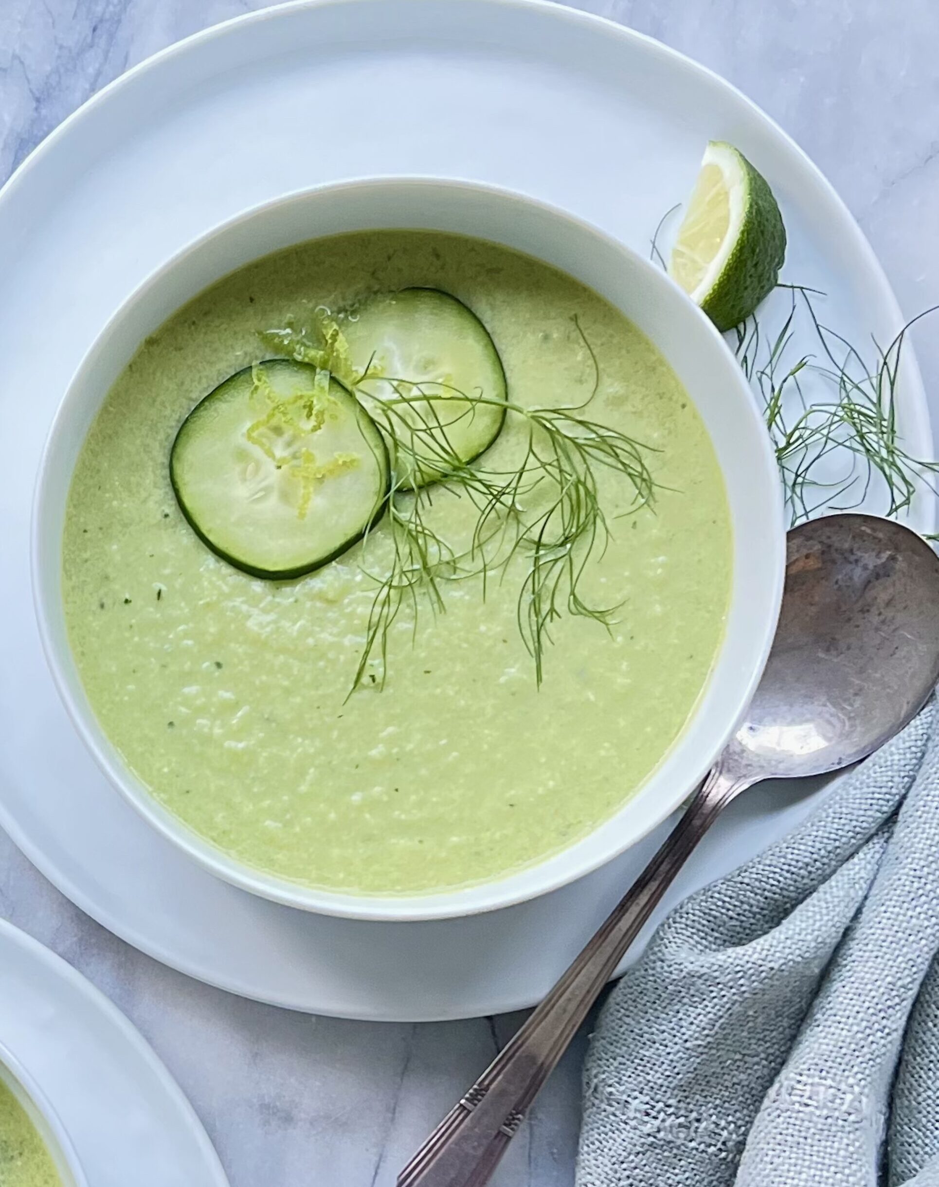 Overhead view of a green gazpacho in a white bowl, garnished by fennel fronds and cucumber slices.