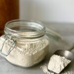 3/4 overhead view of a glass jar of oat flour and to the side if a silver spoon with a spoonful of the flour.