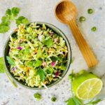 Overhead view of a bowl of the lime cilantro relish on a white background with a spoon resting on the side and a cut lime