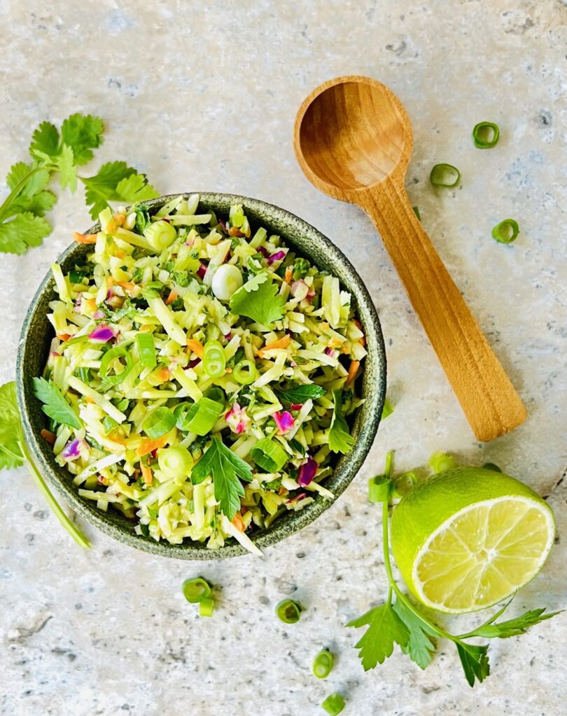 Overhead view of a bowl of the lime cilantro relish on a white background with a spoon resting on the side and a cut lime