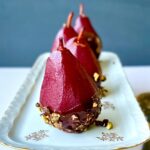 Side view of 4 poached pears, the color of red wine, on a white dish with chocolate and pistachios along the bottom.