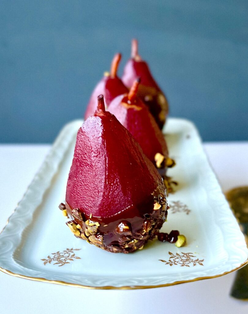 Side view of 4 poached pears, the color of red wine, on a white dish with chocolate and pistachios along the bottom.