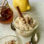 3/4 Overhead shot of a glass bowl with rice pudding with a cinnamon stick in it and cinnamon sprinkled on top. IN the background are a jar of honey, a lemon.