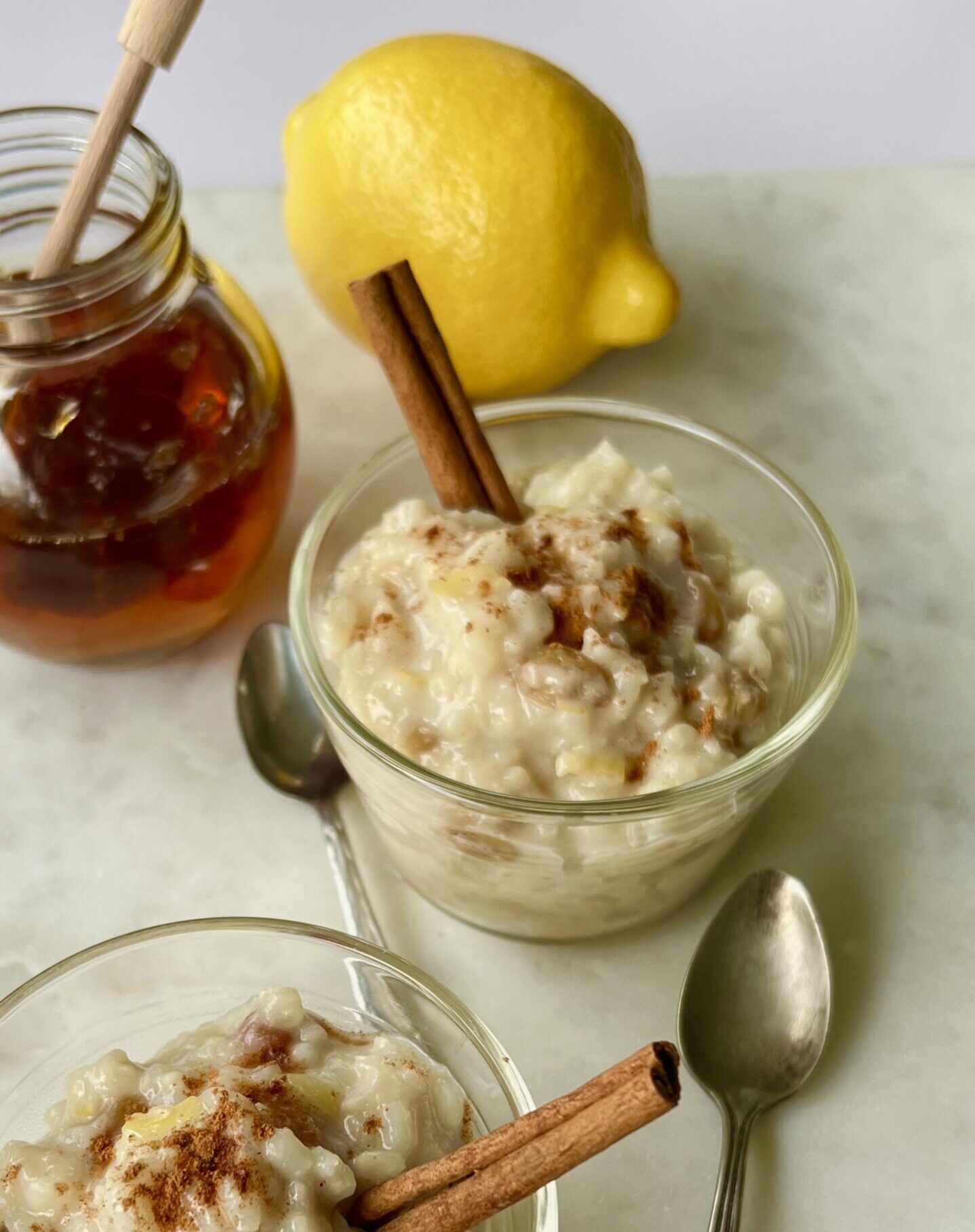 3/4 Overhead shot of a glass bowl with rice pudding with a cinnamon stick in it and cinnamon sprinkled on top. IN the background are a jar of honey, a lemon.