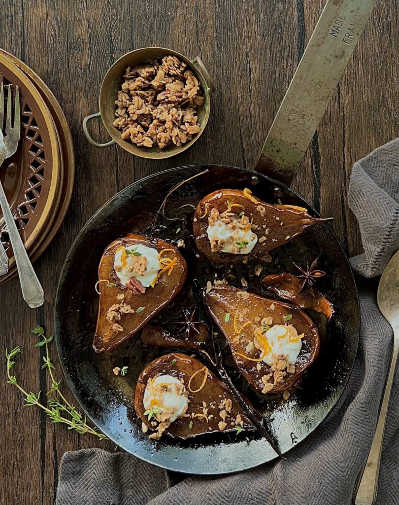 Overhead view of 4 roasted pears in an iron pot on a wood table. The pears have Greek honey and granola on them.